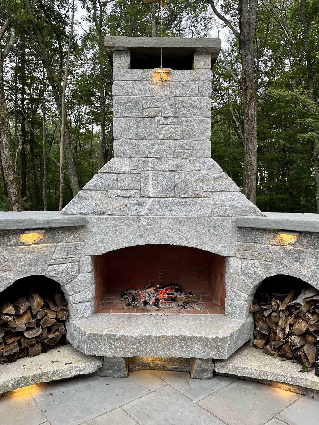 Granite outdoor fireplace with a fire lit and firewood stacked in stone alcoves on each side