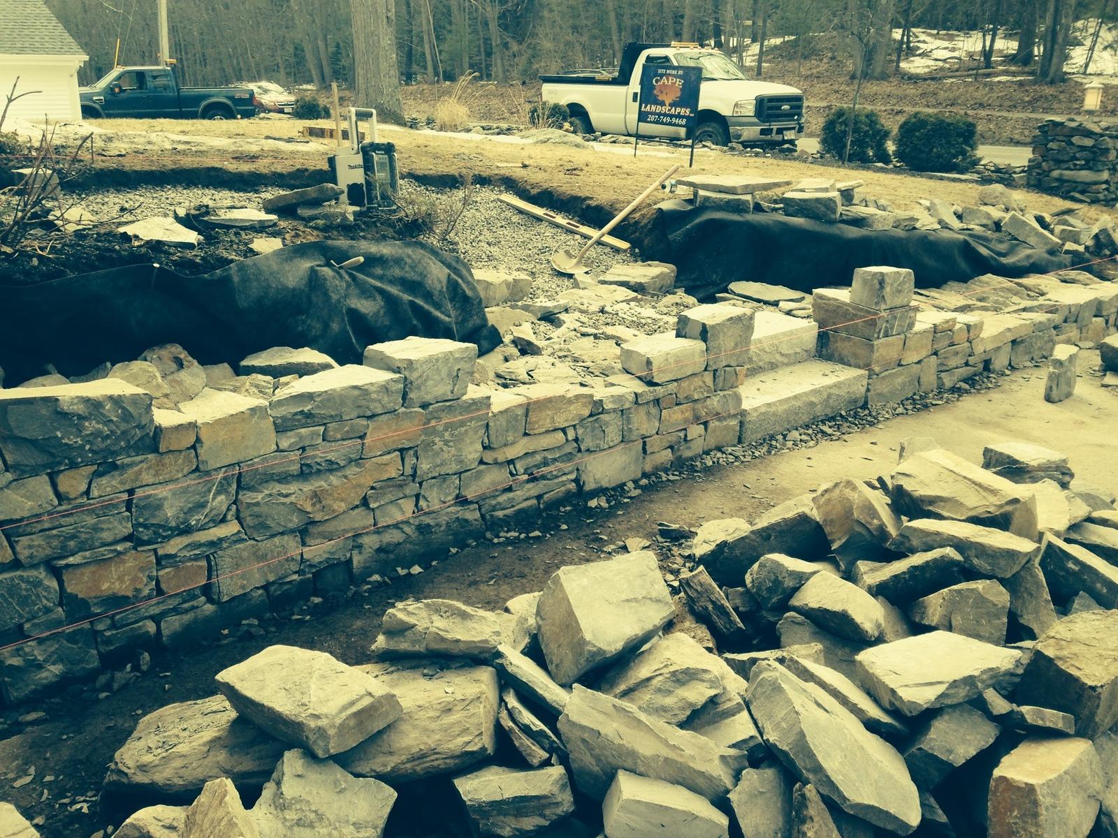 Retaining wall under construction with stone piled nearby and a work truck parked beyond